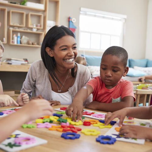 Young Black teacher smiling while a diverse group of children learn with her at a communal desk